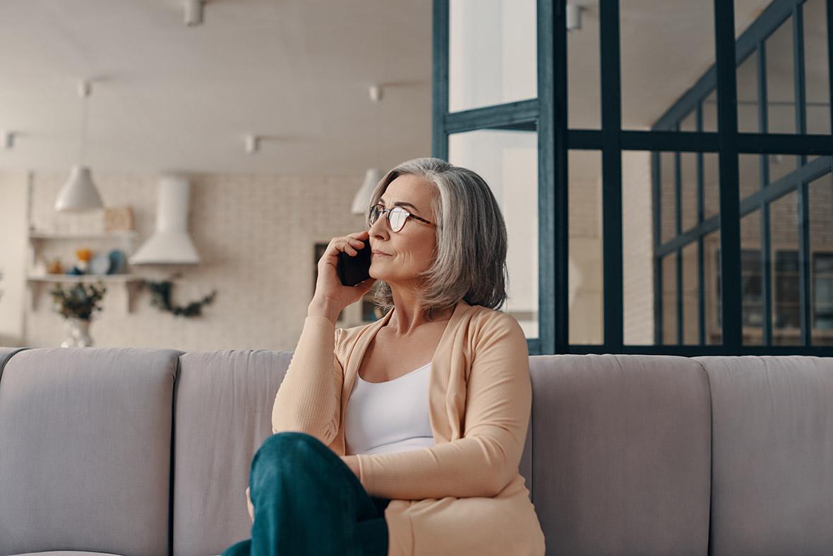 Mature woman in casual clothing talking on phone while sitting on the sofa at home